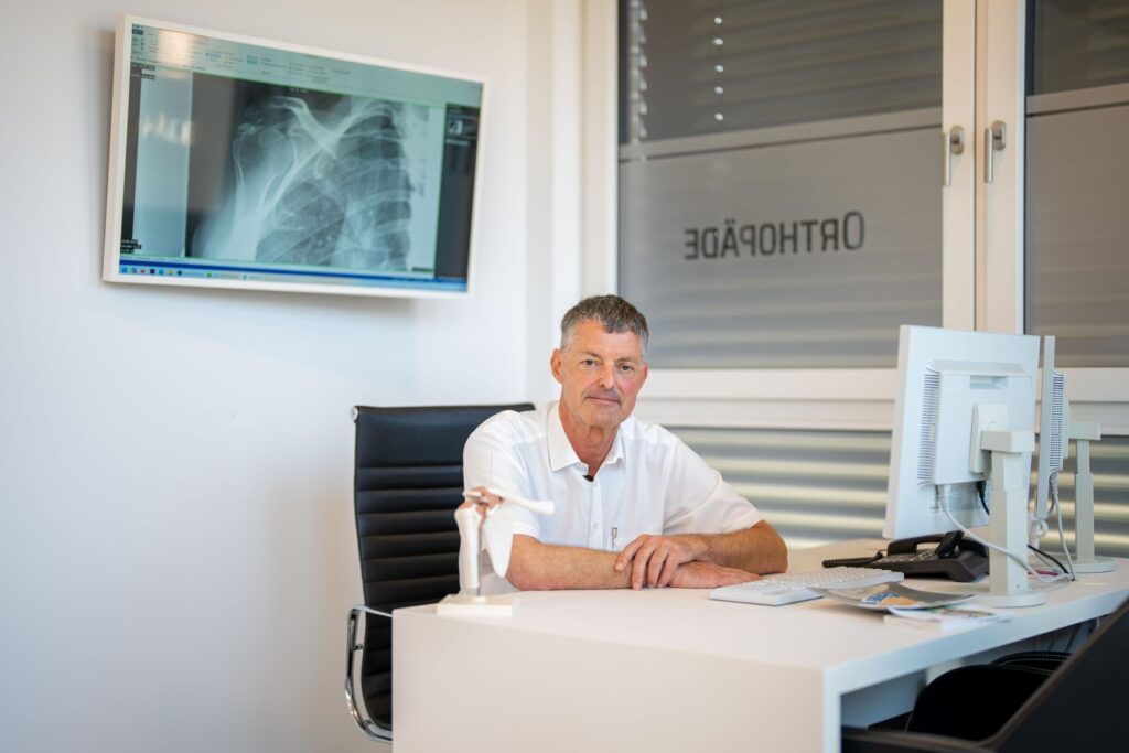 Dr. Knut Bär an seinem Arbeitsplatz, im Hintergrund ein Röntgenbild der Schulter auf dem Monitor